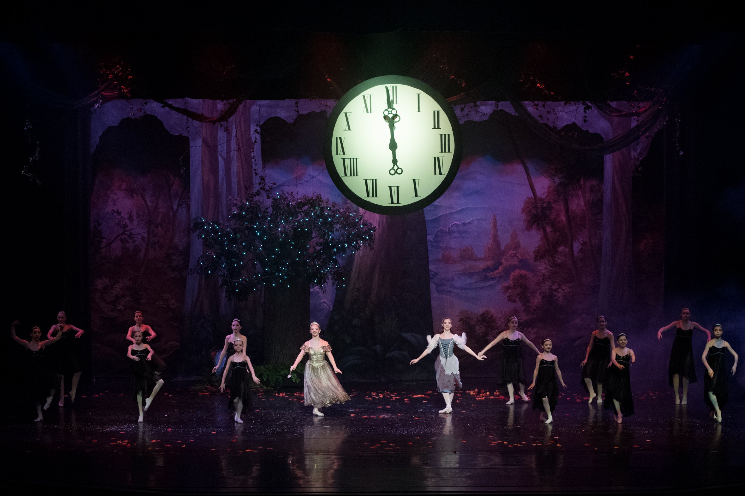 A ballet stage with dancers performing in front of a forest backdrop, featuring a large clock showing midnight and decorated with trees and foliage.