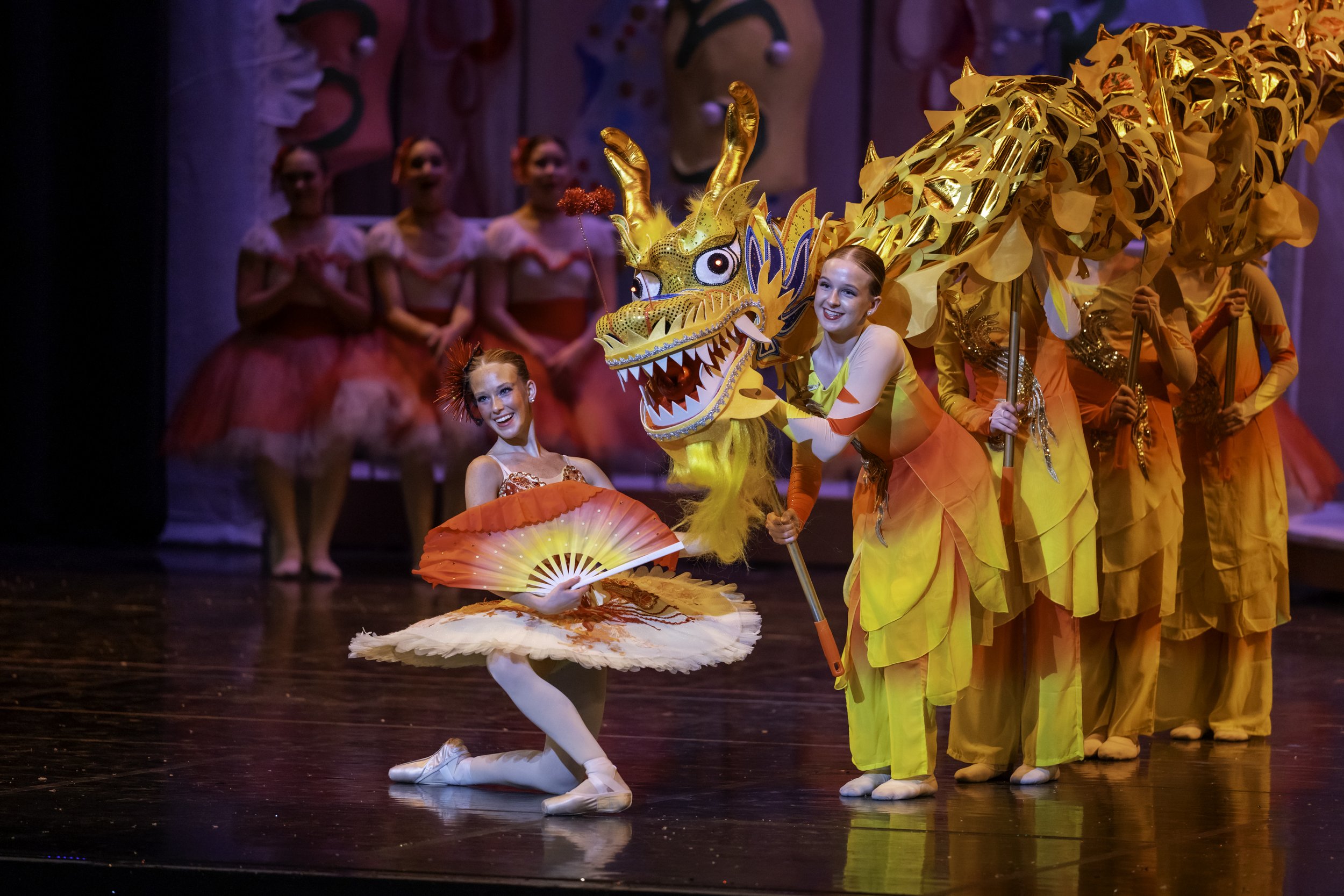 Ballet dancers performing a Chinese New Year themed dance with a girl in a pink and white tutu holding a red folding fan, and another girl holding a colorful dragon prop, with other dancers in yellow costumes and dragon heads in the background.