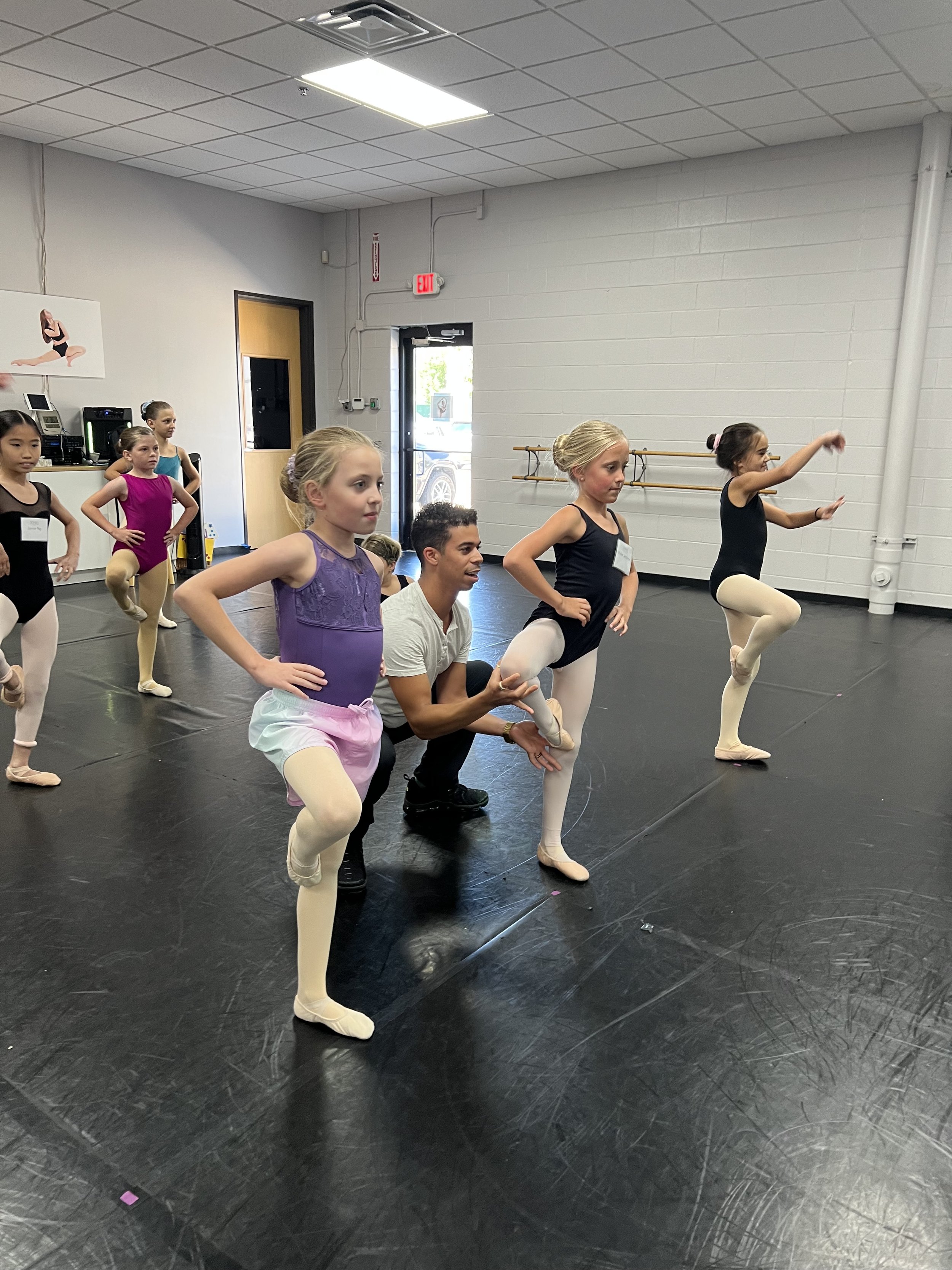 Ballet class with young girls practicing ballet moves in a dance studio, instructor assisting one of the girls.