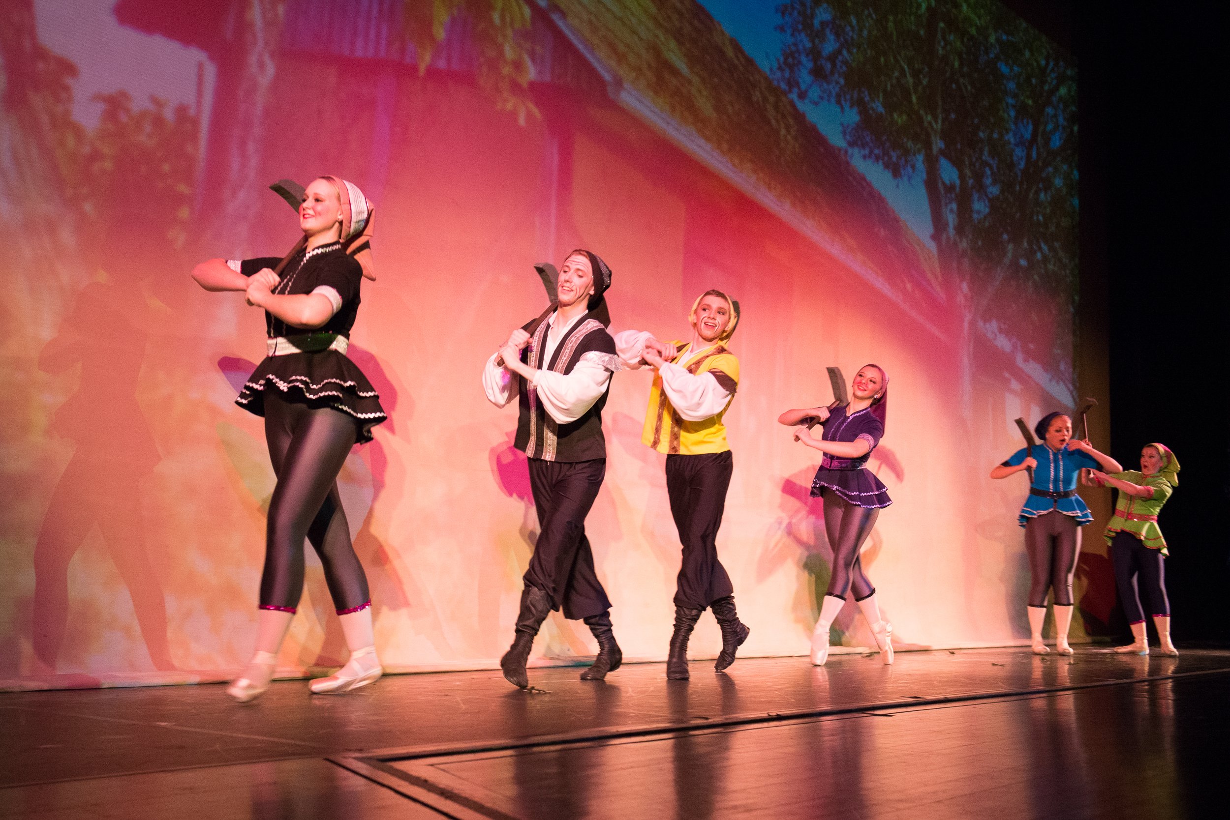Dancers in colorful costumes performing on stage with a scenic backdrop. They are smiling and holding arm positions as part of a choreographed dance.