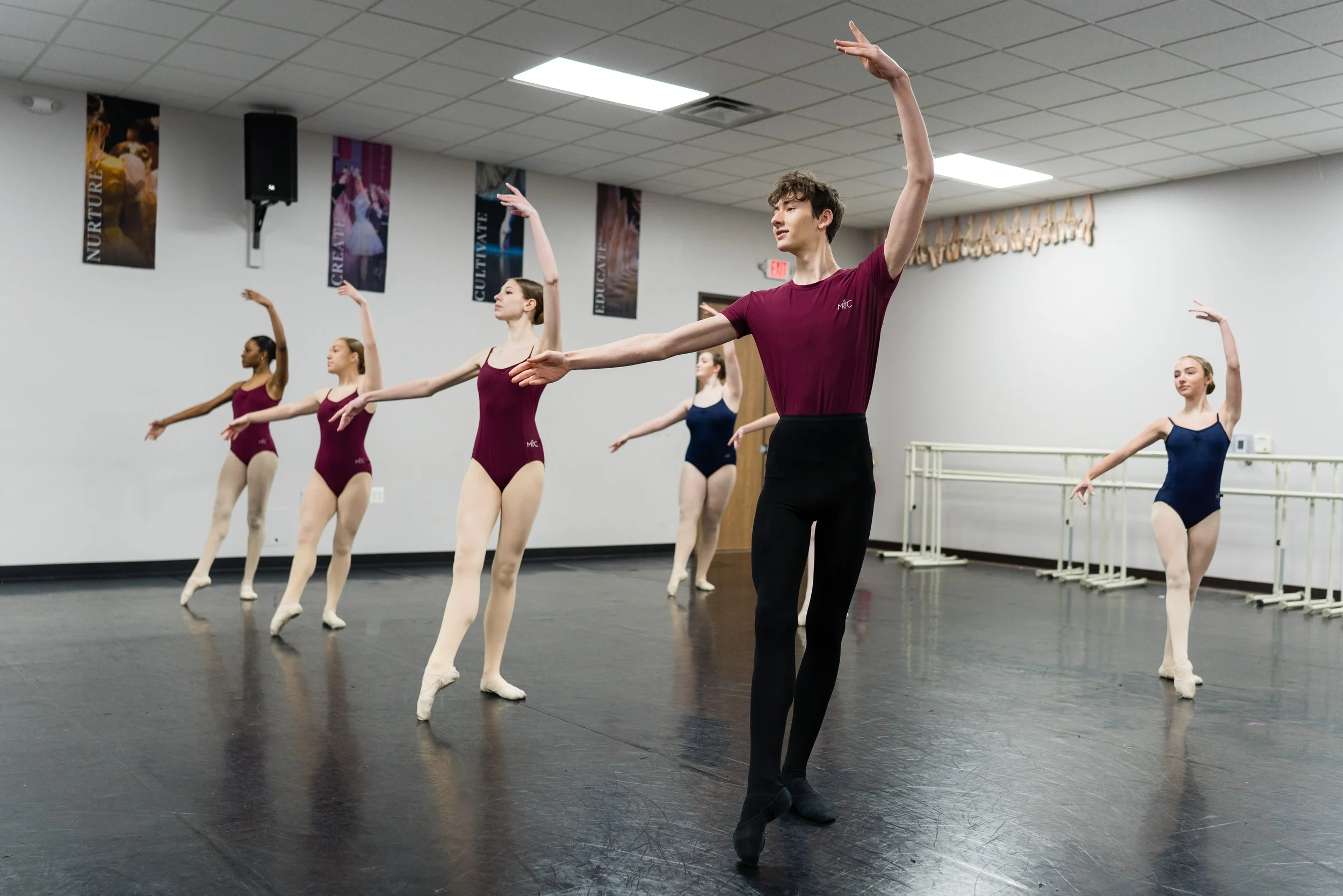 Ballet students in practice at a dance studio, wearing leotards and tights, practicing ballet movements under the guidance of an instructor.