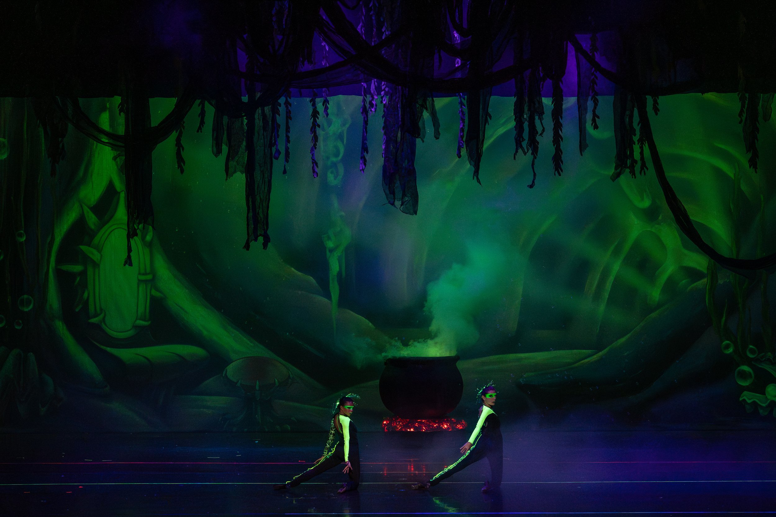 Two performers in black and green costumes with visors perform a dance on stage in a dark, theatrical setting with a mystical forest backdrop. A large cauldron or pot with green smoke rises above it in the background, creating a fantasy or magical at