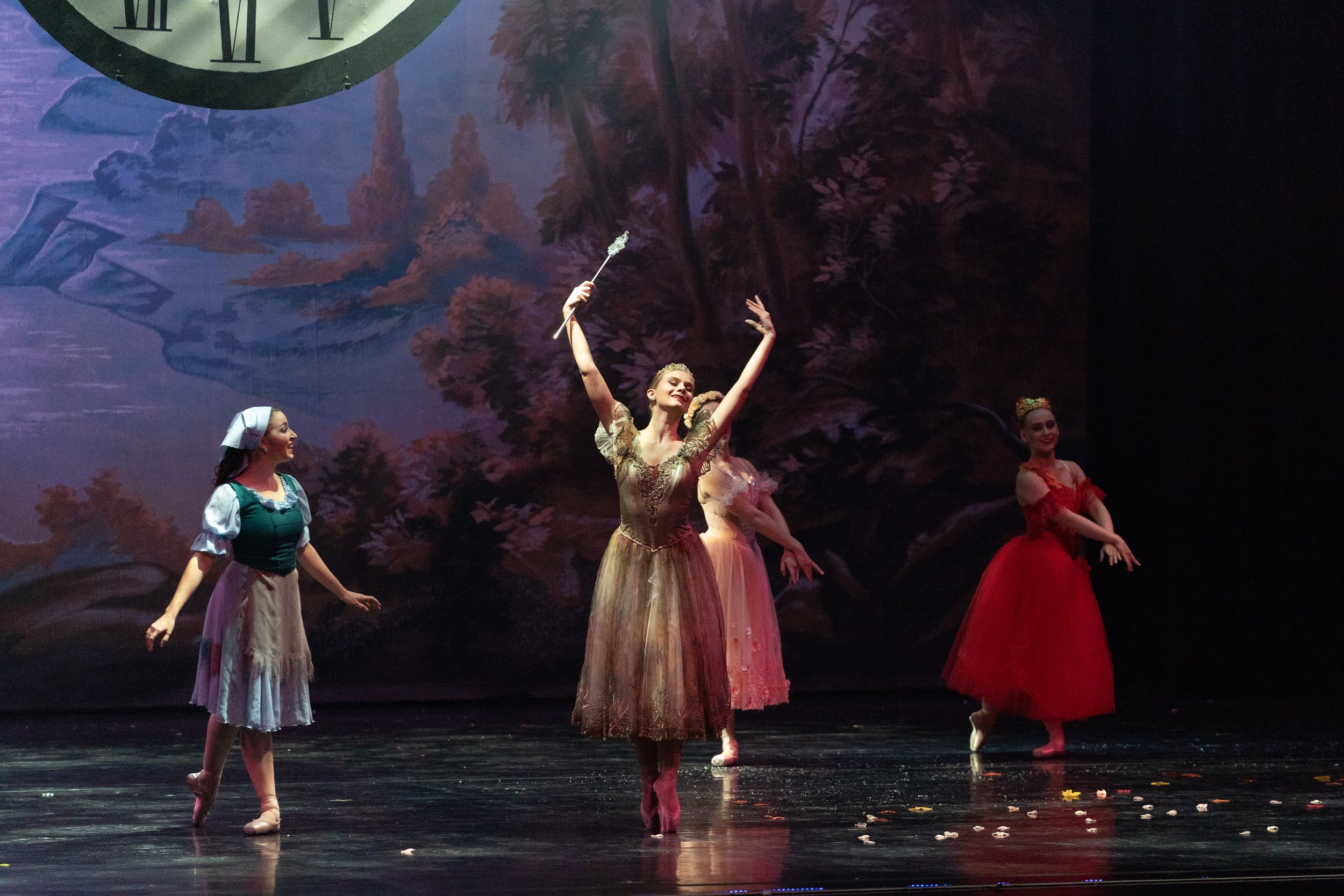 Four ballet dancers performing on stage with a scenic backdrop of a forest and river, one dancer in center holding a wand, others standing with arms extended at various angles.