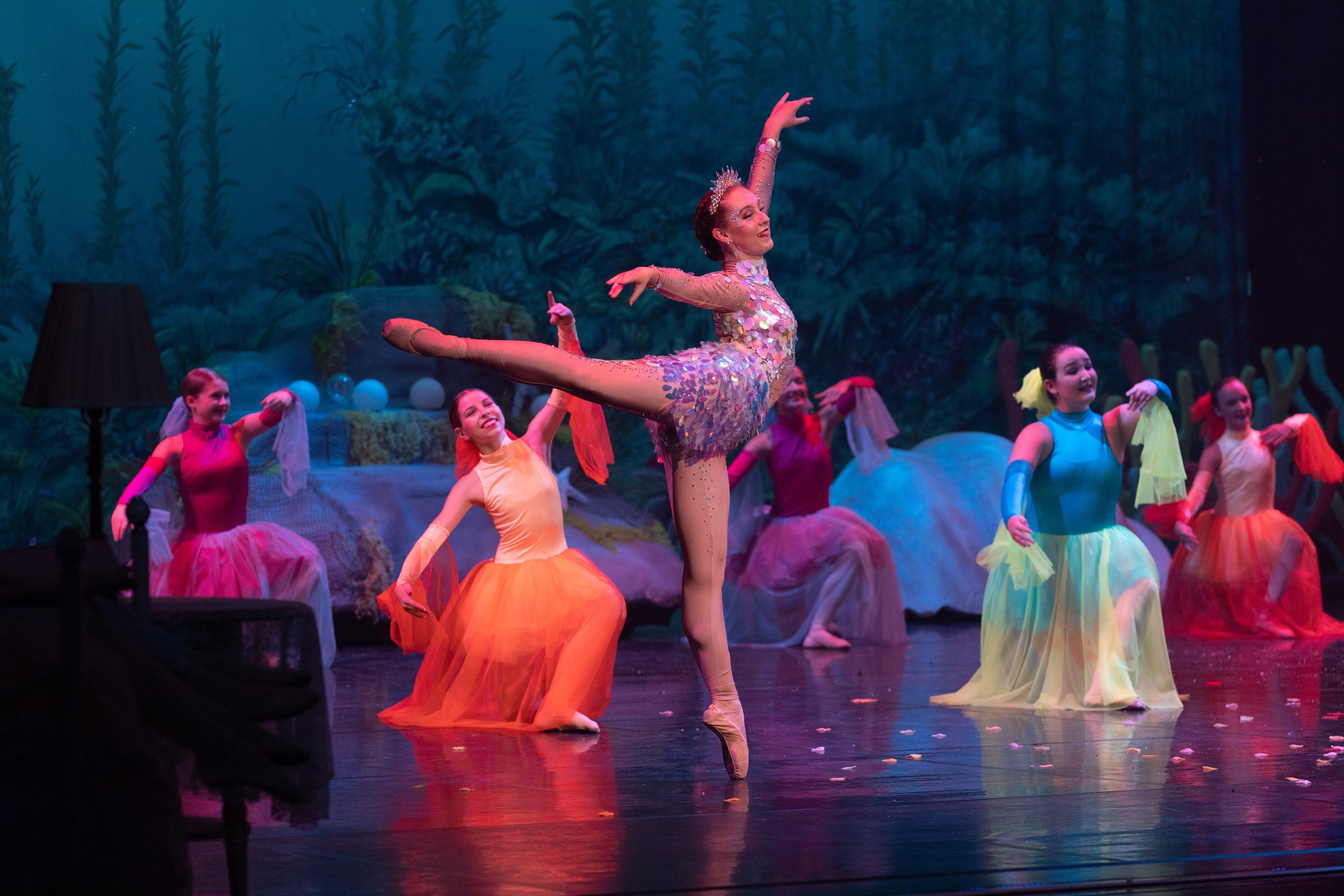 Ballet performance with a lead dancer in a glittery costume and colorful tutu, balancing on point with one leg extended, surrounded by fellow dancers in vibrant costumes on stage with a forest backdrop.