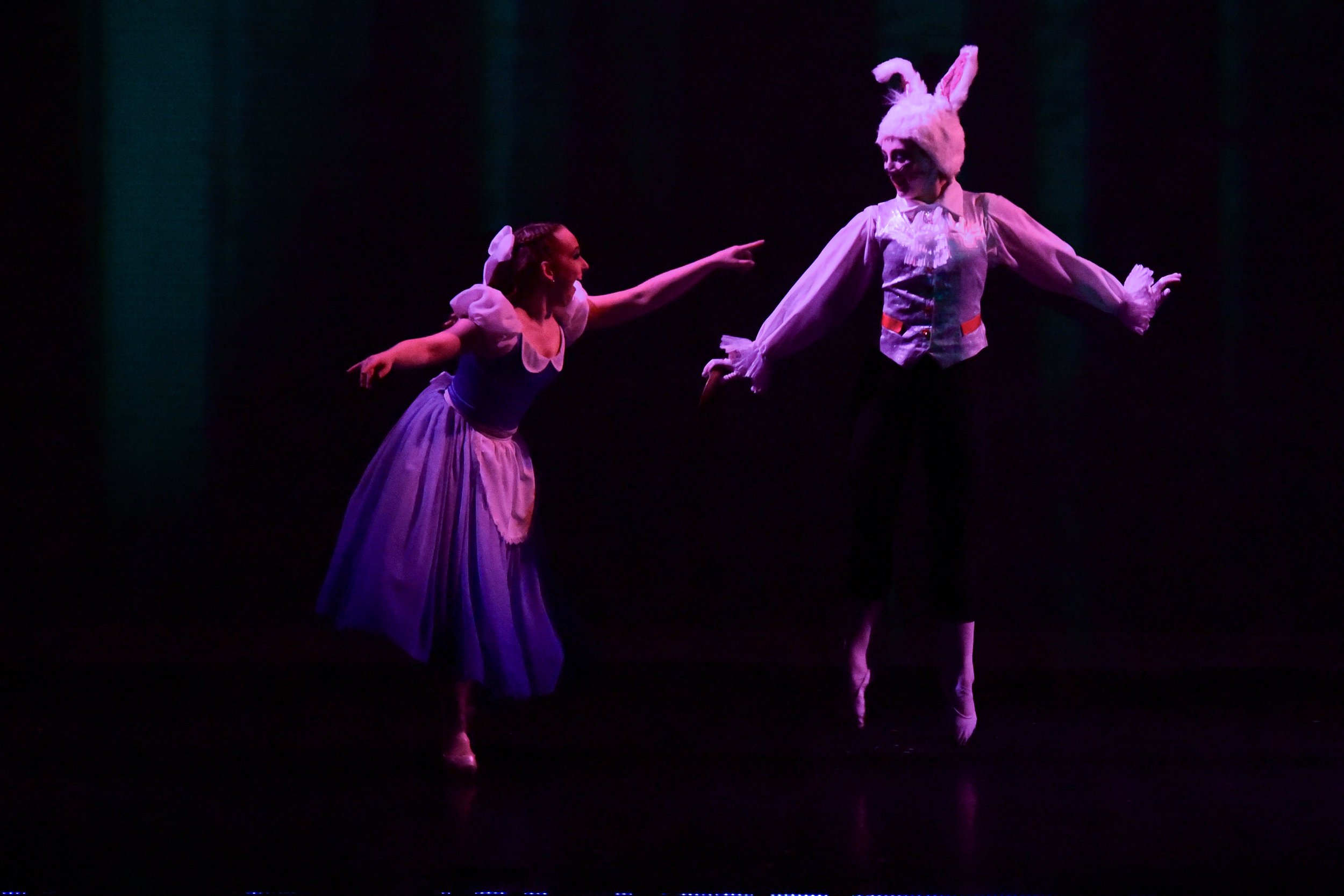 Two ballet dancers performing on stage, one dressed as a woman and the other as a rabbit, in a theatrical scene with dark background and colored lighting.