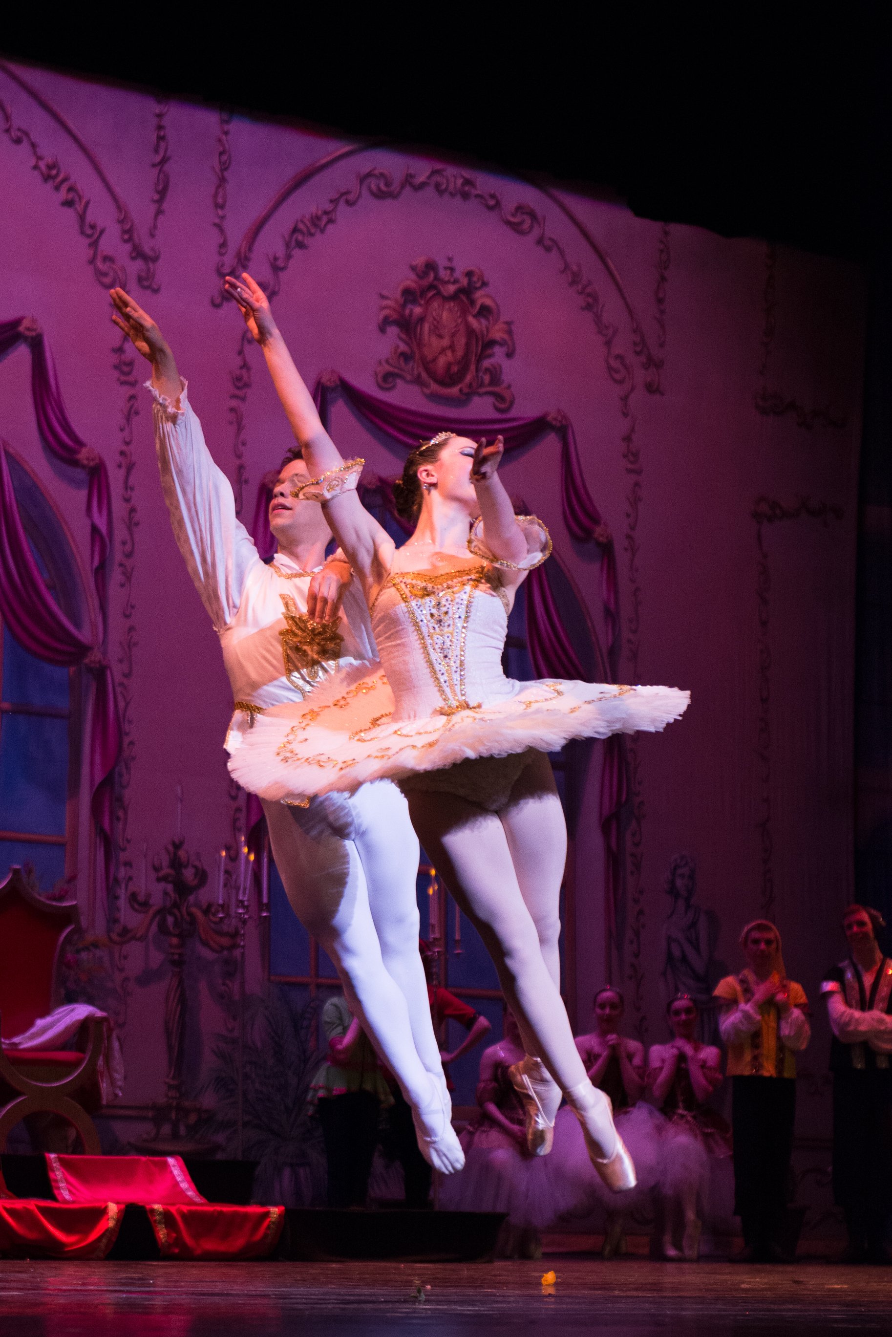Two ballet dancers in white and gold costumes perform on stage, with one dancer mid-air and the other behind her. The backdrop features a pink and purple ornate interior design with spectators in period costumes watching.
