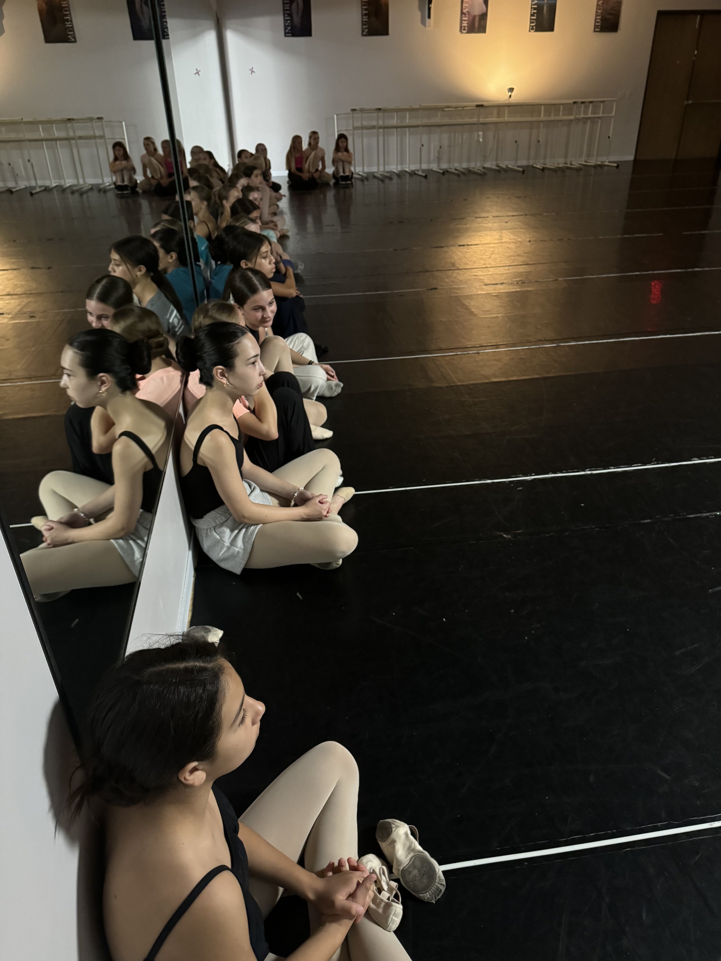 Ballet dancers sitting against a wall in a dance studio, with a large mirror reflecting their image and dance posters on the wall.