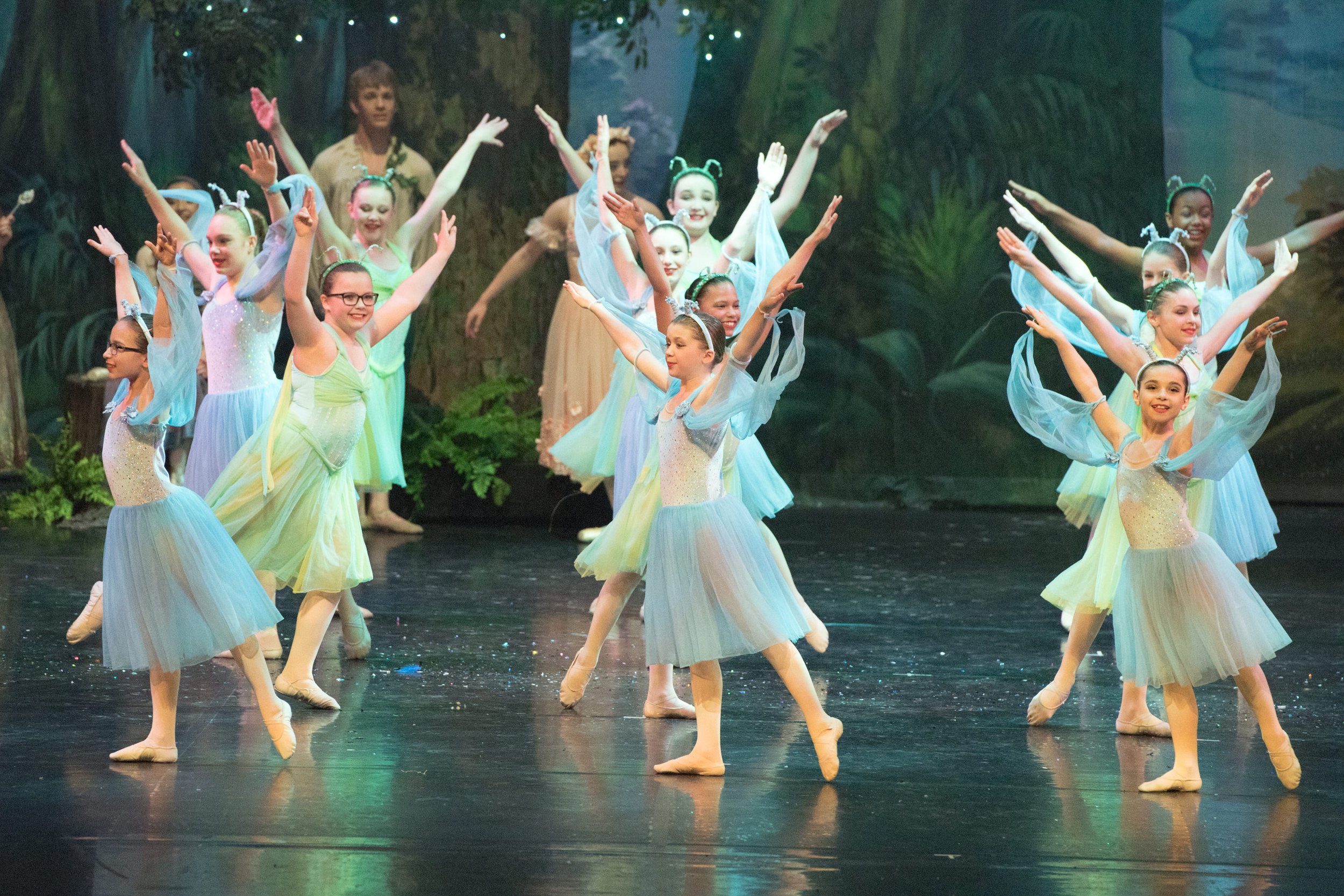 Group of young girls performing a ballet dance on stage, dressed in pastel-colored tutus with tiaras, in a forest-themed backdrop.