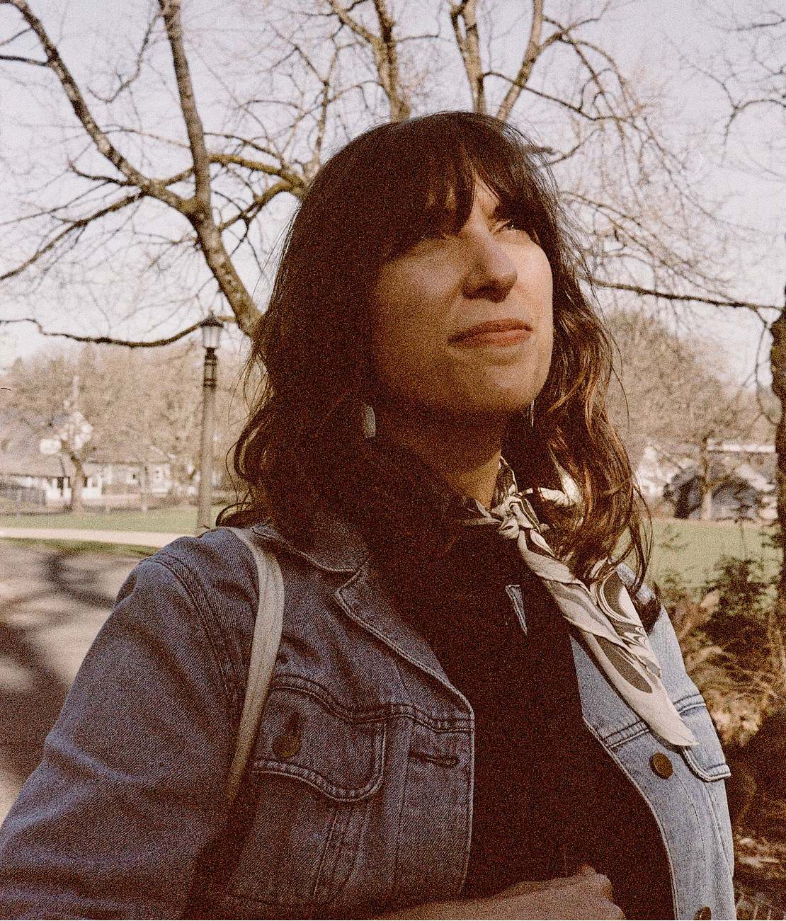 A young woman with dark wavy hair and a patterned scarf around her neck, outdoors in a park with bare trees and a cloudy sky, captured with film photography.