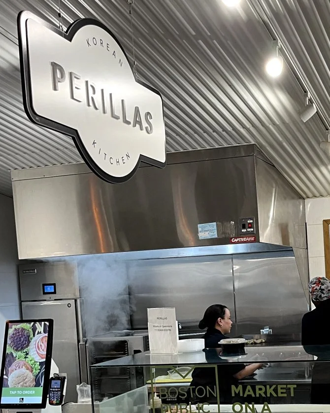 Interior view of a Korean kitchen restaurant named Perillas, with a large metallic ventilation hood, a sign overhead, and a steam-filled cooking area where a woman and a customer are present.