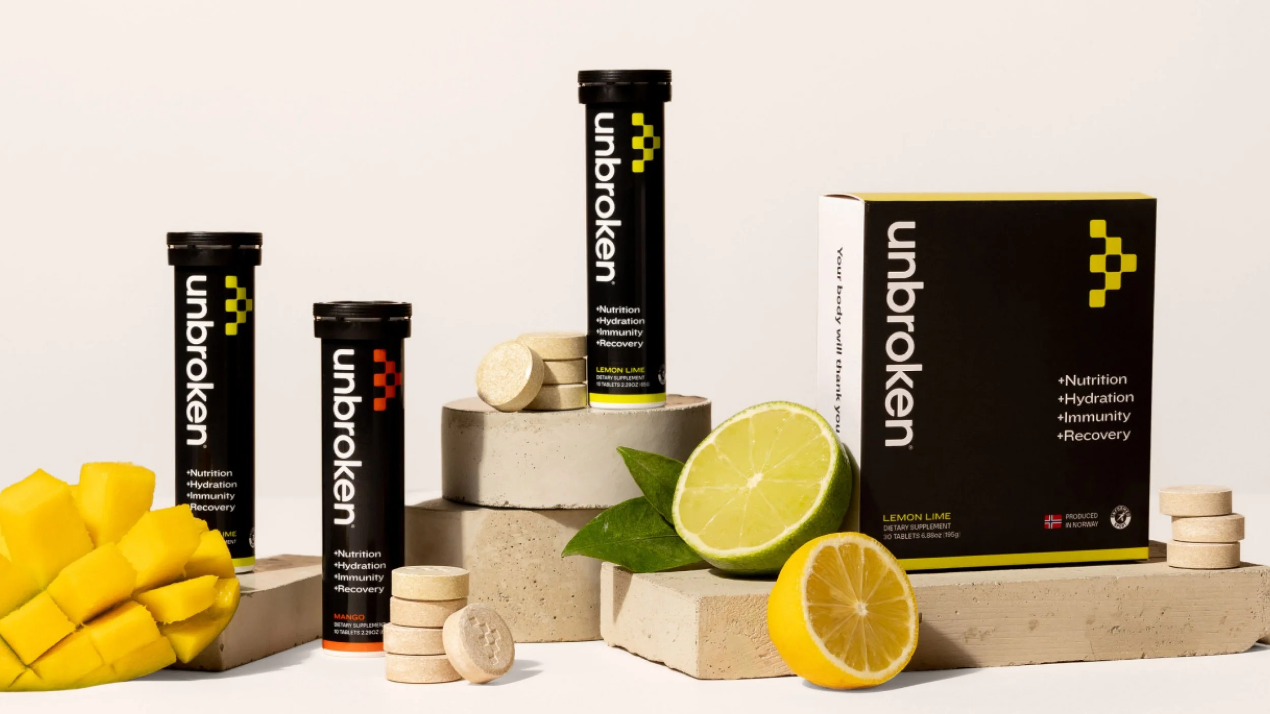 Display of Unbroken dietary supplement tubes in front of sliced lemon, lime, and mango, on a cream-colored background. The packaging is black with yellow and orange accents, and there are tablets arranged around them.