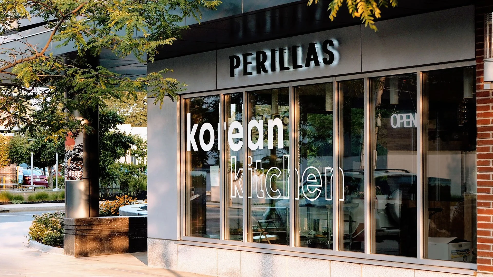 Exterior of Perillas Korean kitchen restaurant with large glass windows, white and black signage, and an 'OPEN' sign visible inside.