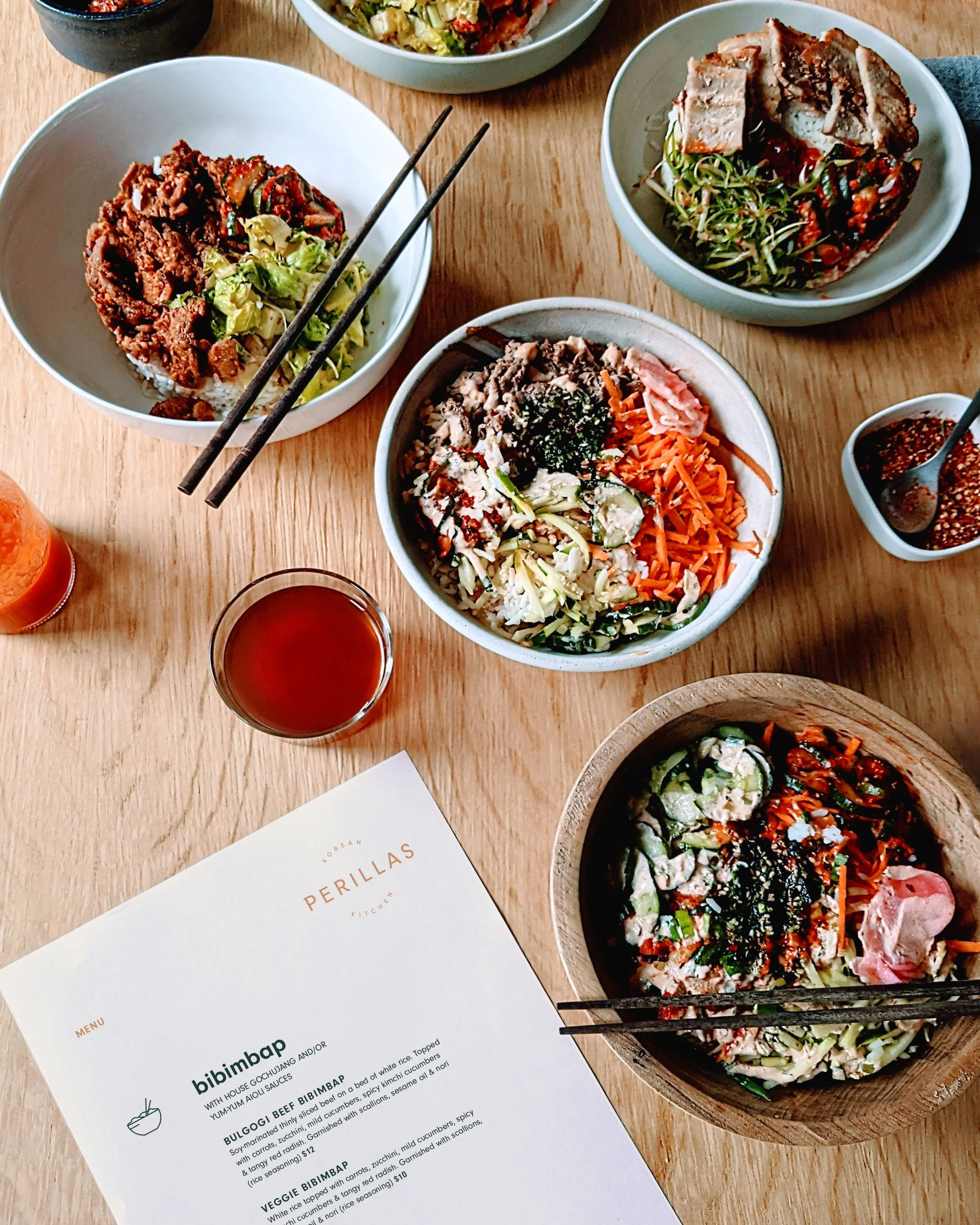 An assortment of Korean dishes on a wooden table, including bibimbap bowls, chopsticks, dipping sauces, a drink, and a menu from Perillas Korean Kitchen.