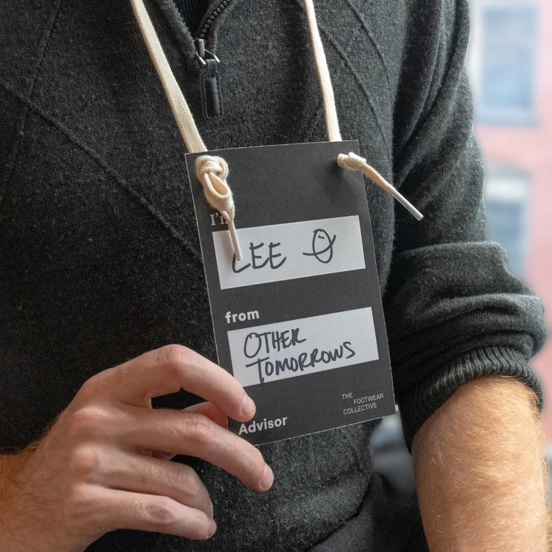 A person wearing a dark gray zip-up hoodie holding a black and white name tag that says LEE, from OTHER TOMORROWS, with a doodle of a face. The person has short hair and the background shows an out-of-focus window.