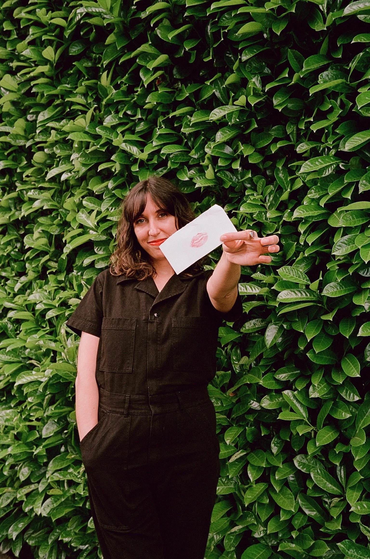 A woman with curly brown hair wearing a black outfit, standing in front of a green leafy hedge, holding a white envelope with a red lipstick kiss stamp towards the camera, captured with film photography.