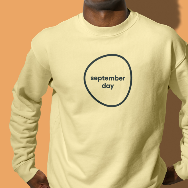Person wearing a light yellow sweatshirt with a black circle around the words 'september day' printed in the center, standing against a two-tone orange and brown background.