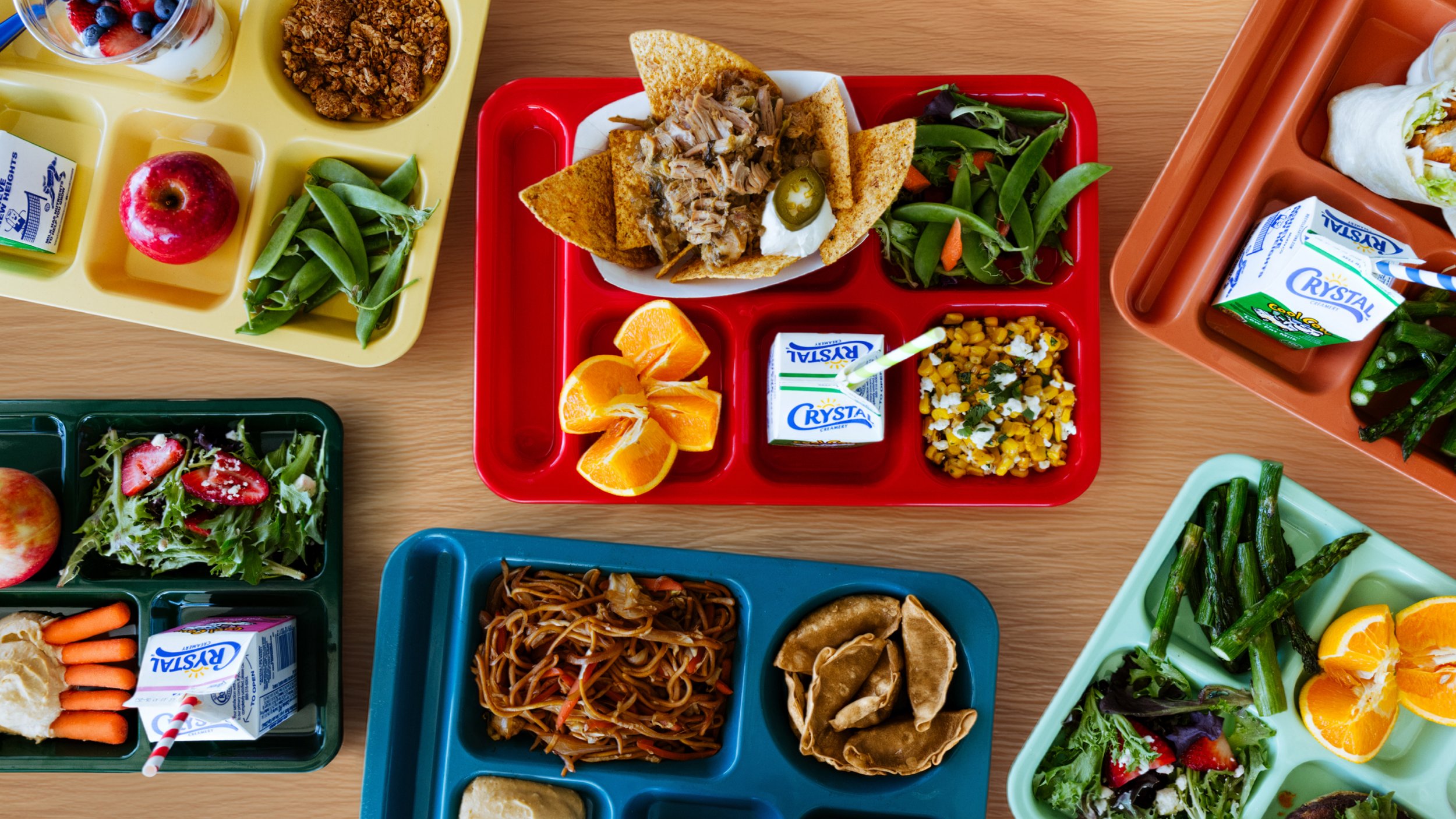 Colorful school lunch trays with various food items including salad, fruit, vegetables, noodles, and drinks.
