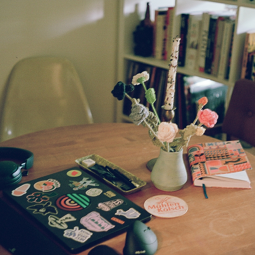 A wooden table with a variety of items including a laptop covered with colorful stickers, a closed notebook, a vase with knitted flowers, a notebook, a pen, a round coaster with text, and headphones. In the background, there's a bookshelf filled with books.