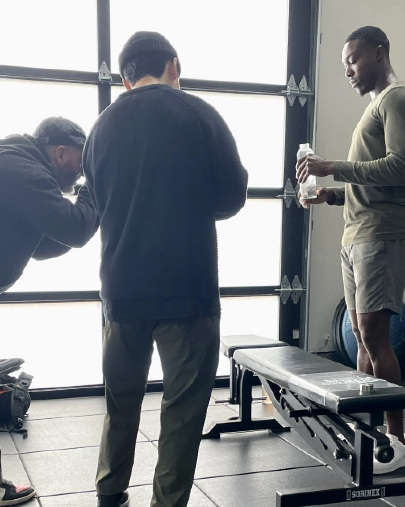 Three people in workout gear in a gym near a large window with frosted glass, one with a water bottle, another adjusting equipment, and one with a beanie looking down.