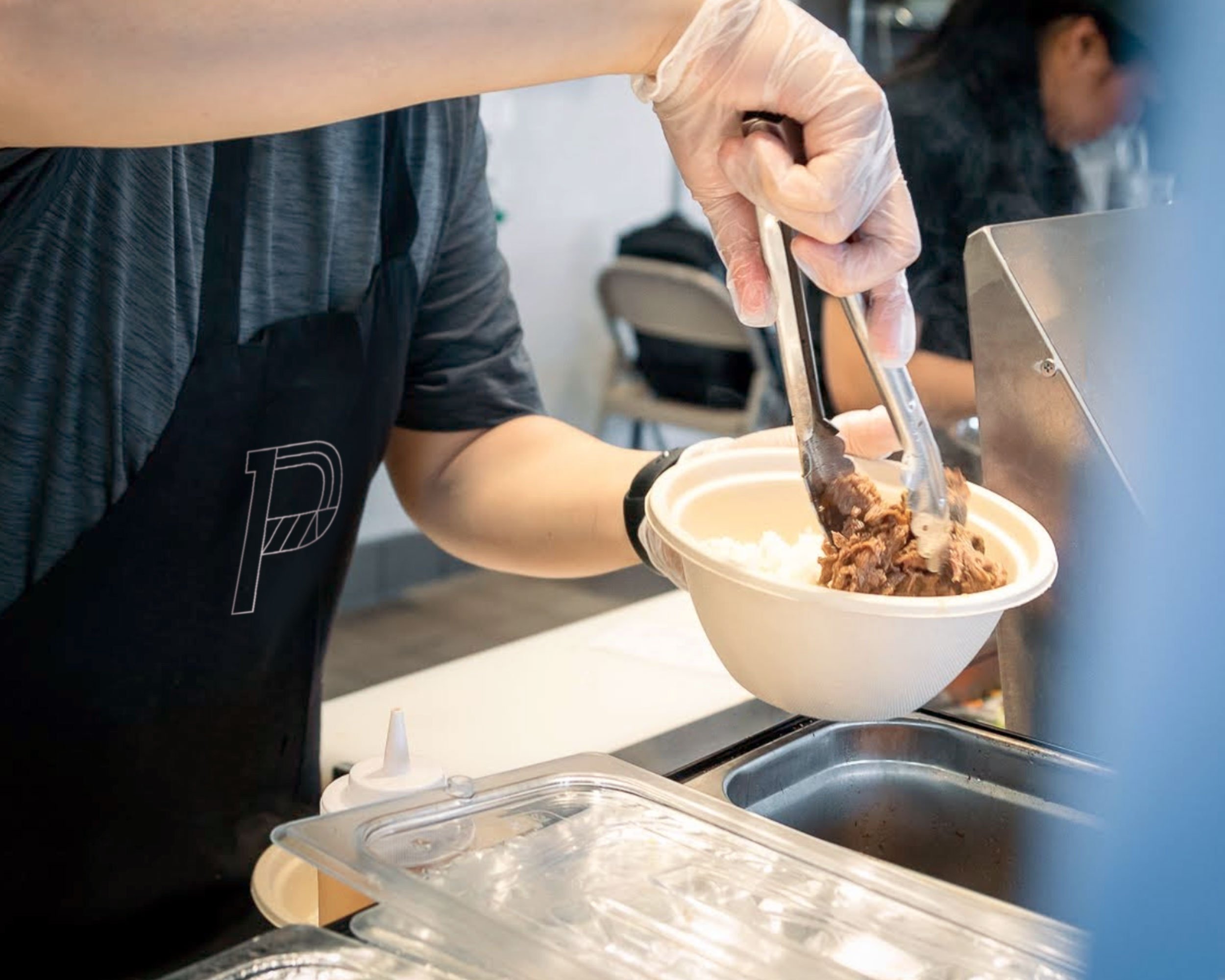 Person in a black apron with a P logo on it and gloves serving beef bulgogi into a bowl with tongs at a food service counter.