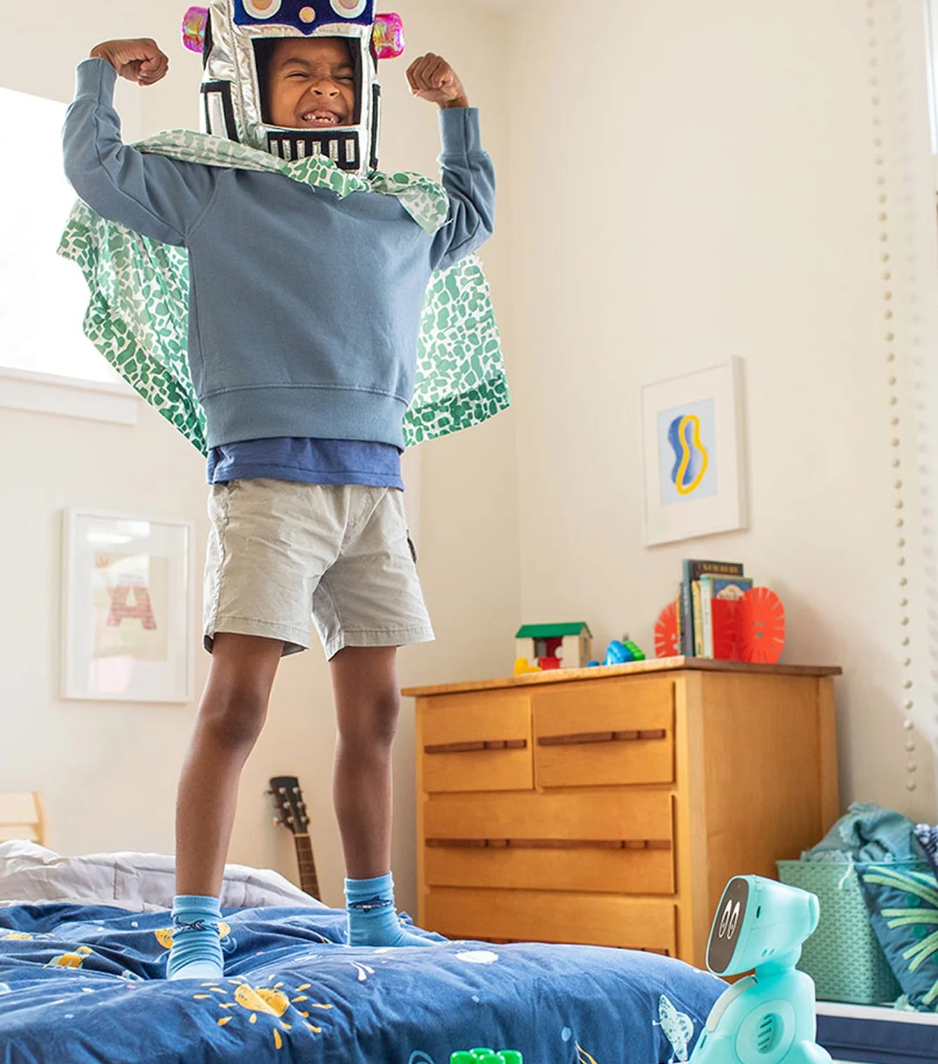 A child standing on a bed wearing a robot costume with a helmet, flexing their arms, and smiling.