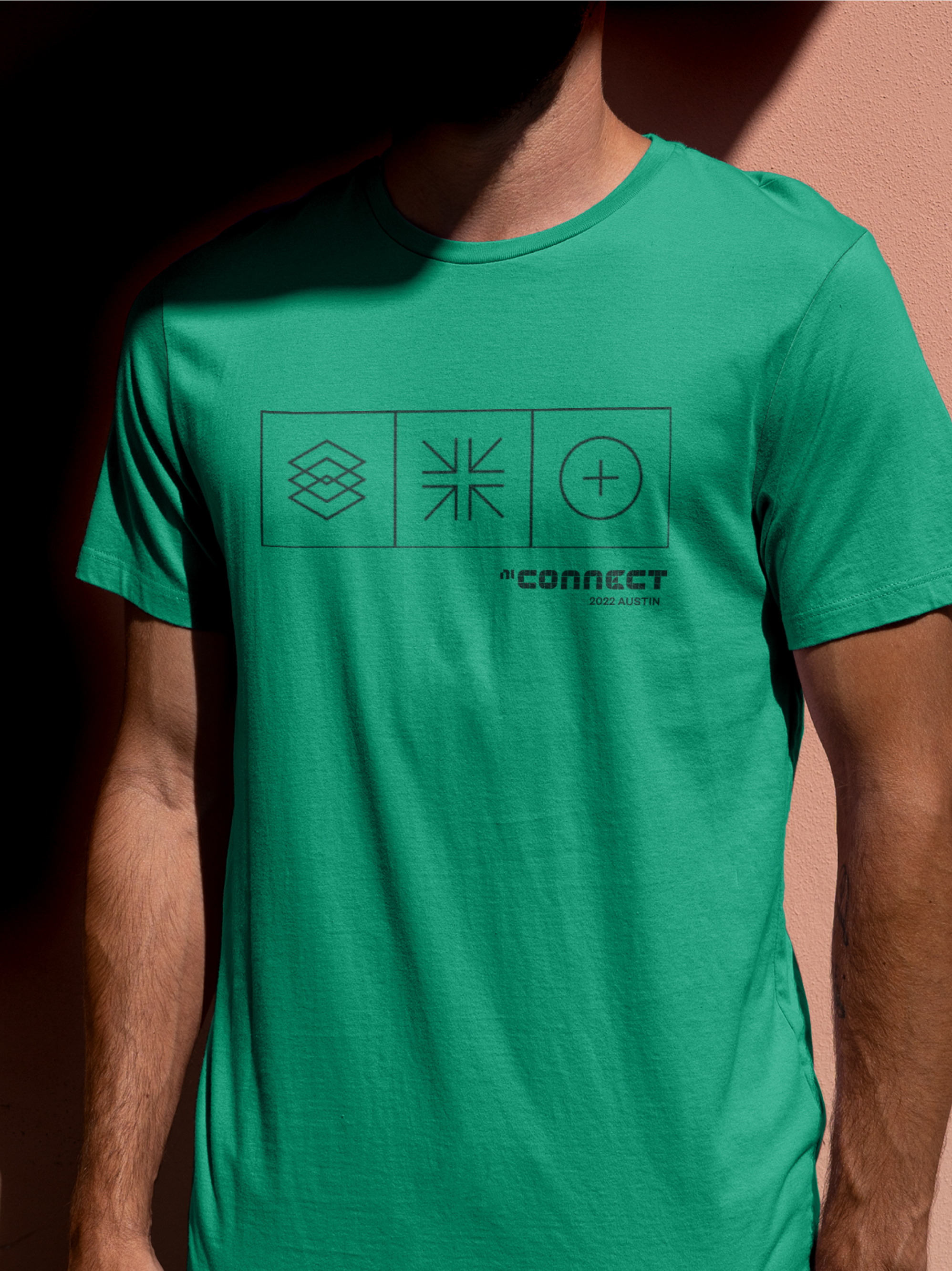 Close-up of a person wearing a green t-shirt with black graphic symbols and text, standing against a pink wall with shadow.