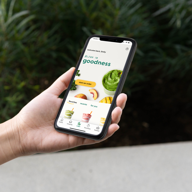 A person holding a smartphone displaying a smoothie app against a blurred green outdoor background.