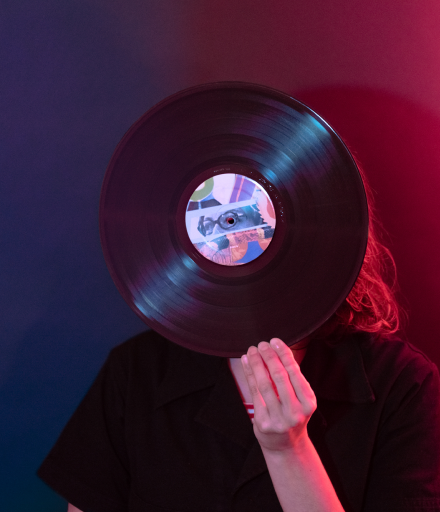 Person holding a vinyl record in front of their face, obscuring their identity, with colorful, gradated lighting in the background.