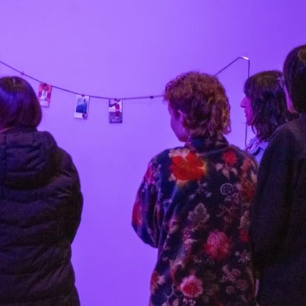 Three people looking at photographs hanging on a string in a dimly lit room with purple lighting.