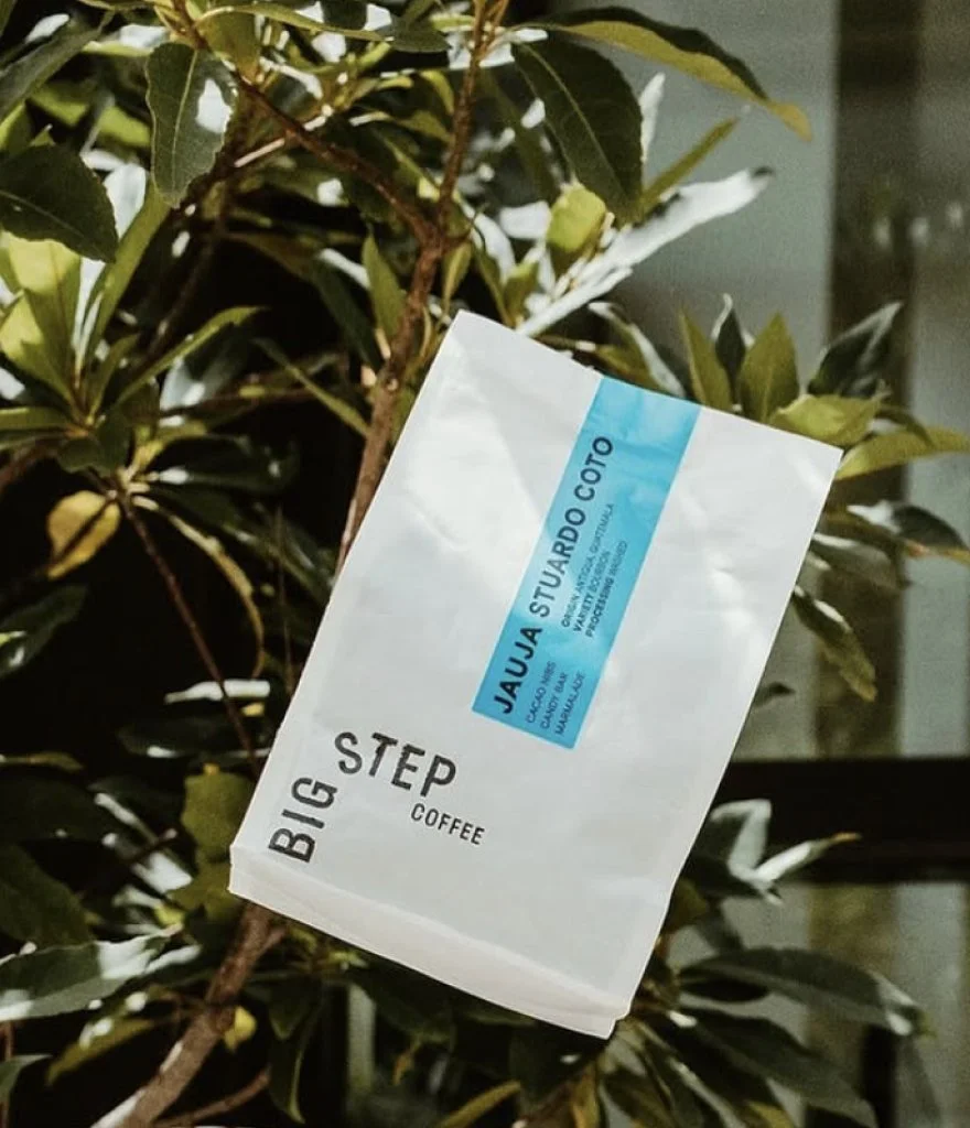 A coffee bag labeled "Big Step Coffee" is being thrown into the air in front of a green leafy plant.