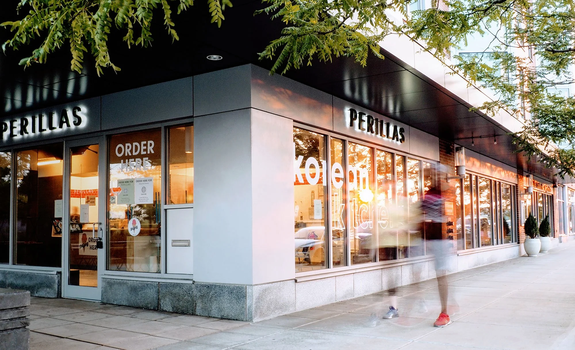 Exterior view of a restaurant named Perillas Korean Kitchen with large glass windows, illuminated signs, and a person walking by during sunset.
