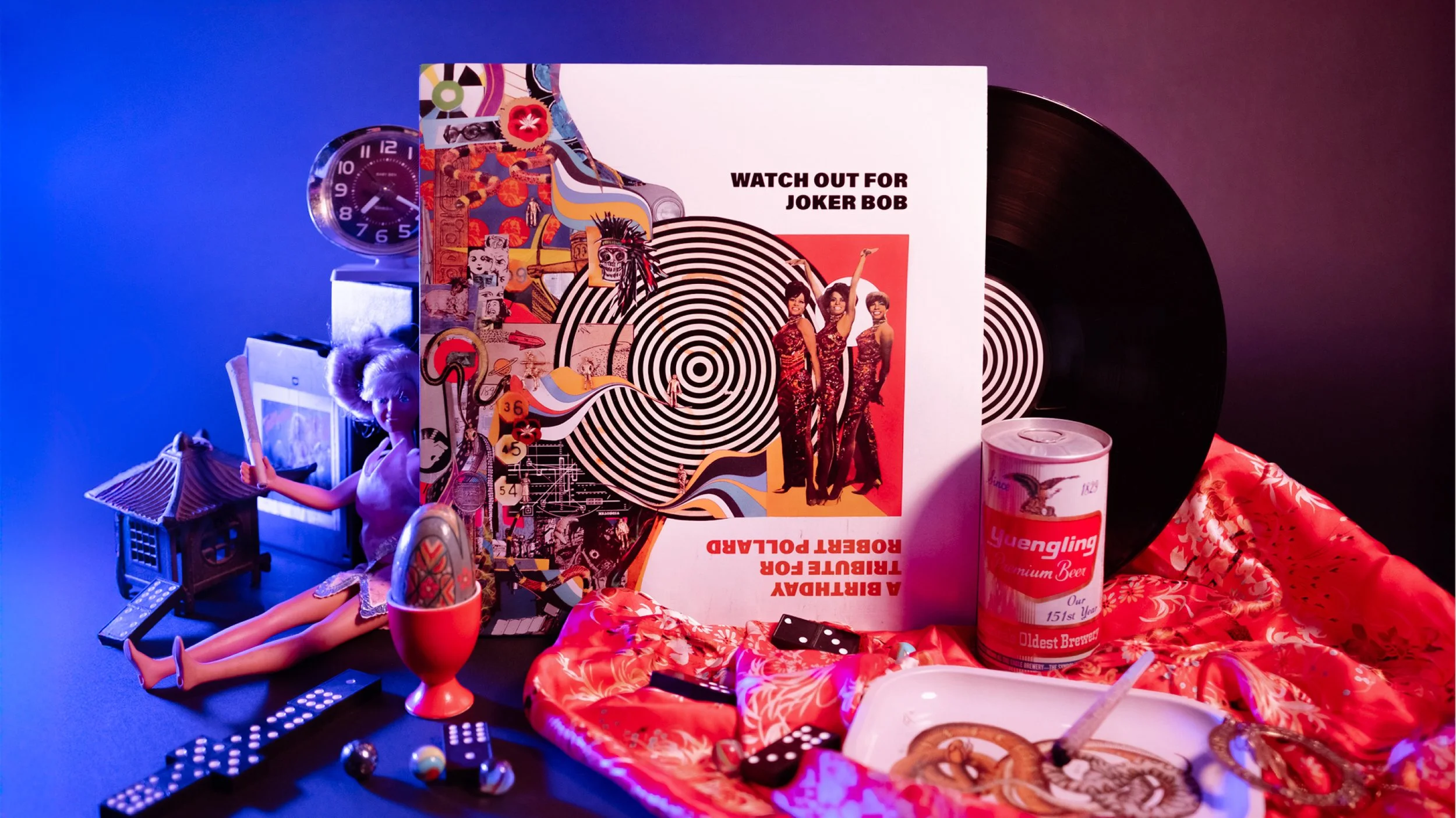 A collection of eclectic objects on a black surface with a blue and purple background, including a large vinyl record, a framed album cover titled "A Birthday Tribute for Joker Bob" with vibrant artwork and three women, a Barbie doll seated on the surface, a toy house, a red cup, a can of Yuengling beer, a red satin cloth, dice, a tray with snacks, and various small trinkets.