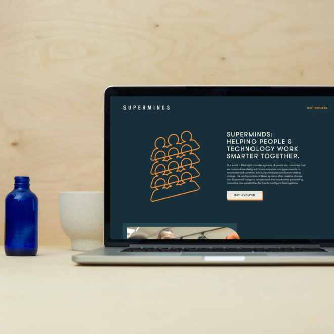 Laptop displaying a website titled 'Superminds' with a graphic of stacked people icons and text about helping people and technology work smarter together, placed on a light wooden desk next to a blue glass bottle and a beige speckled cup.