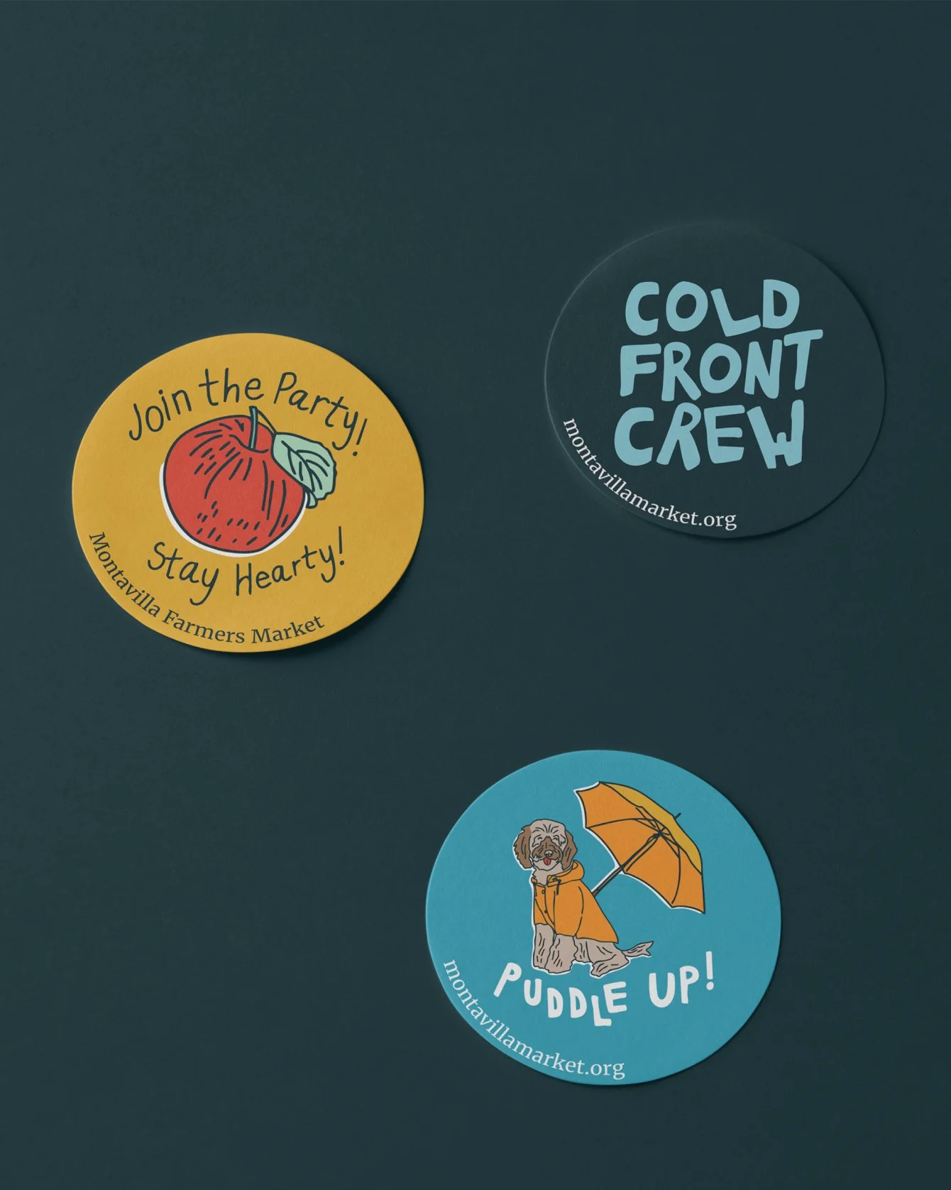 Three circular stickers on a dark background. The yellow sticker features a red apple with green leaf and the texts 'Join the Party! Stay Hearty! Mountvila Farmers Market'. The dark blue sticker has the text 'COLD FRONT CREW' and 'montavilamarket.org'. The light blue sticker shows a dog with a raincoat and umbrella with the text 'PUDDLE UP!' and 'mountavilamarket.org'.