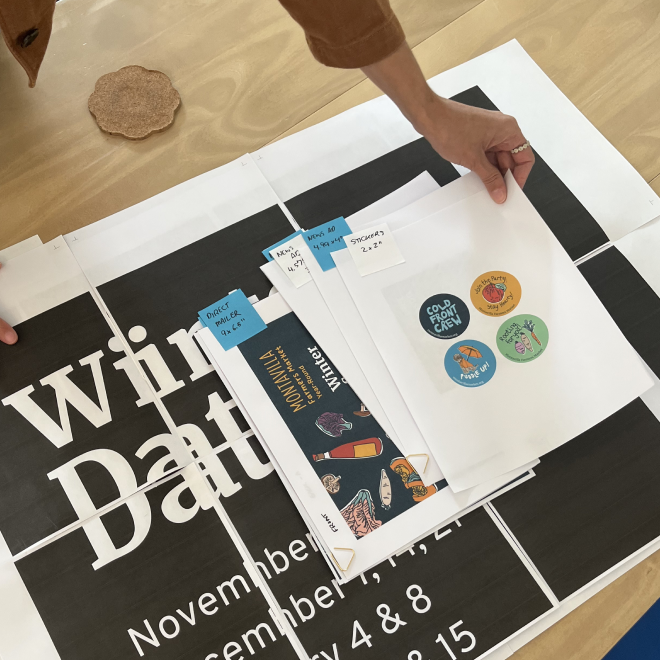 Person organizing paper materials on a table, which include event posters, sticker sheets, and printed papers being reviewed.