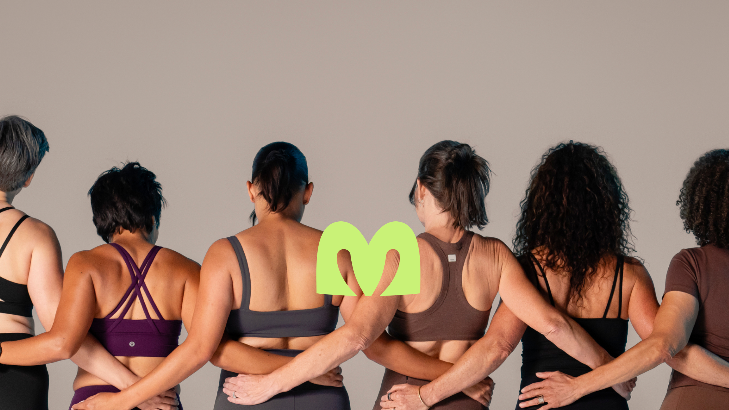 Seven women standing in a line with arms around each other's waists against a plain grey background and an "M"-shaped logo overlaying in neon yellow.