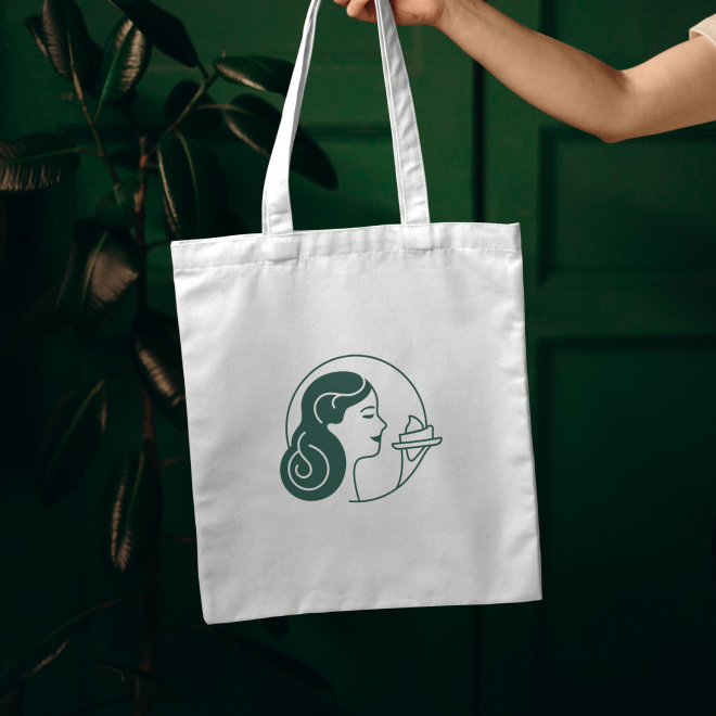 White tote bag with a line drawing of a woman holding a plate near her face, against a green wall background.