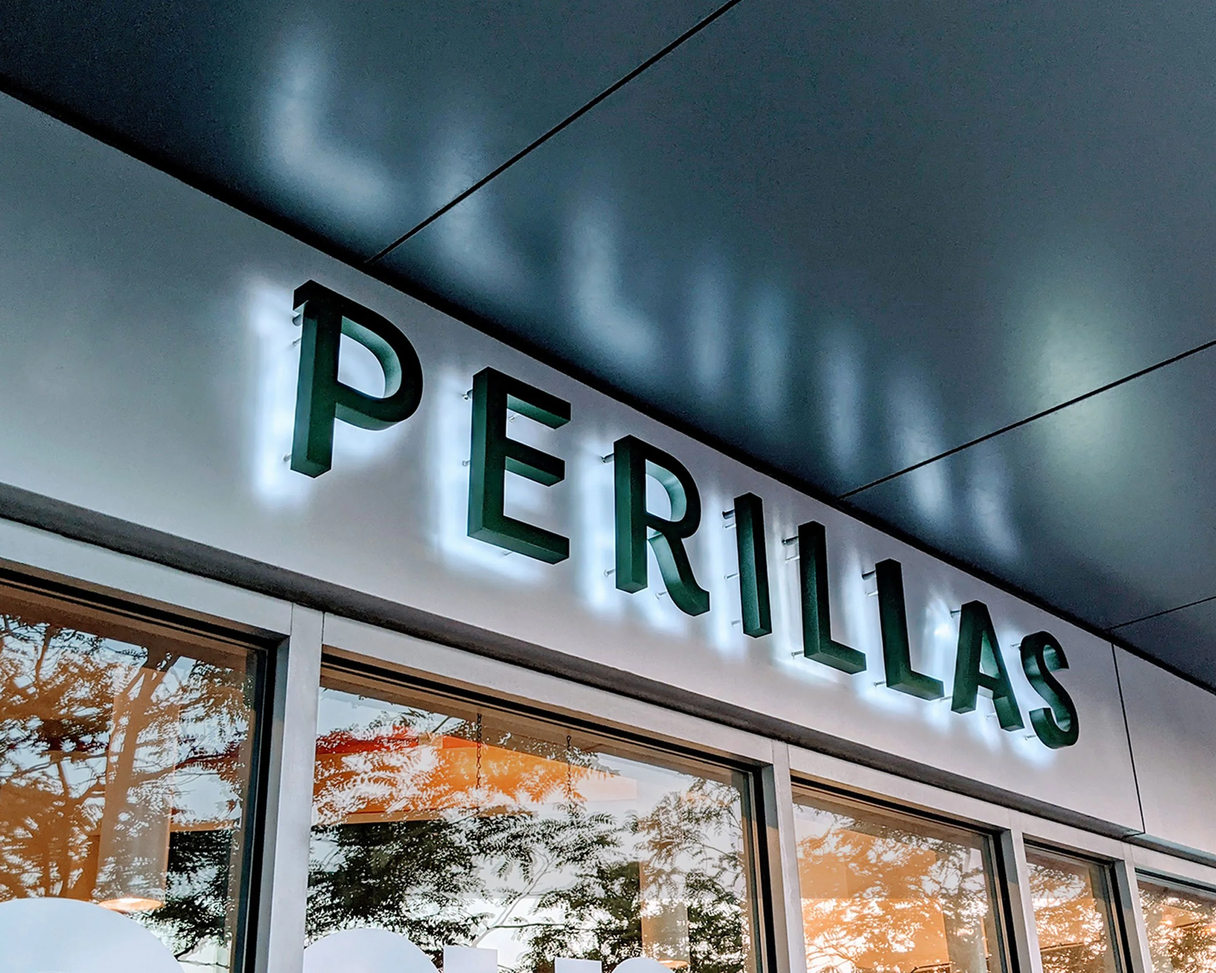 Sign displaying the word 'PERILLAS' above a window reflecting orange lights and trees.