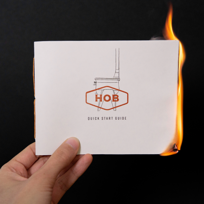 A hand holding a booklet titled 'HOB Quick Start Guide' that is on fire on the right side, with a black background.