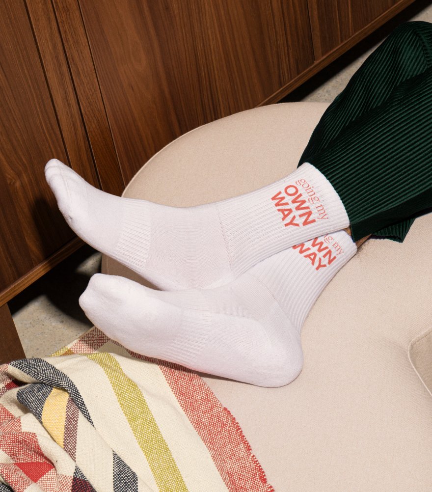 Person's legs and feet resting on a beige chair, wearing white socks with red text and green pants, with a multicolored striped blanket nearby.