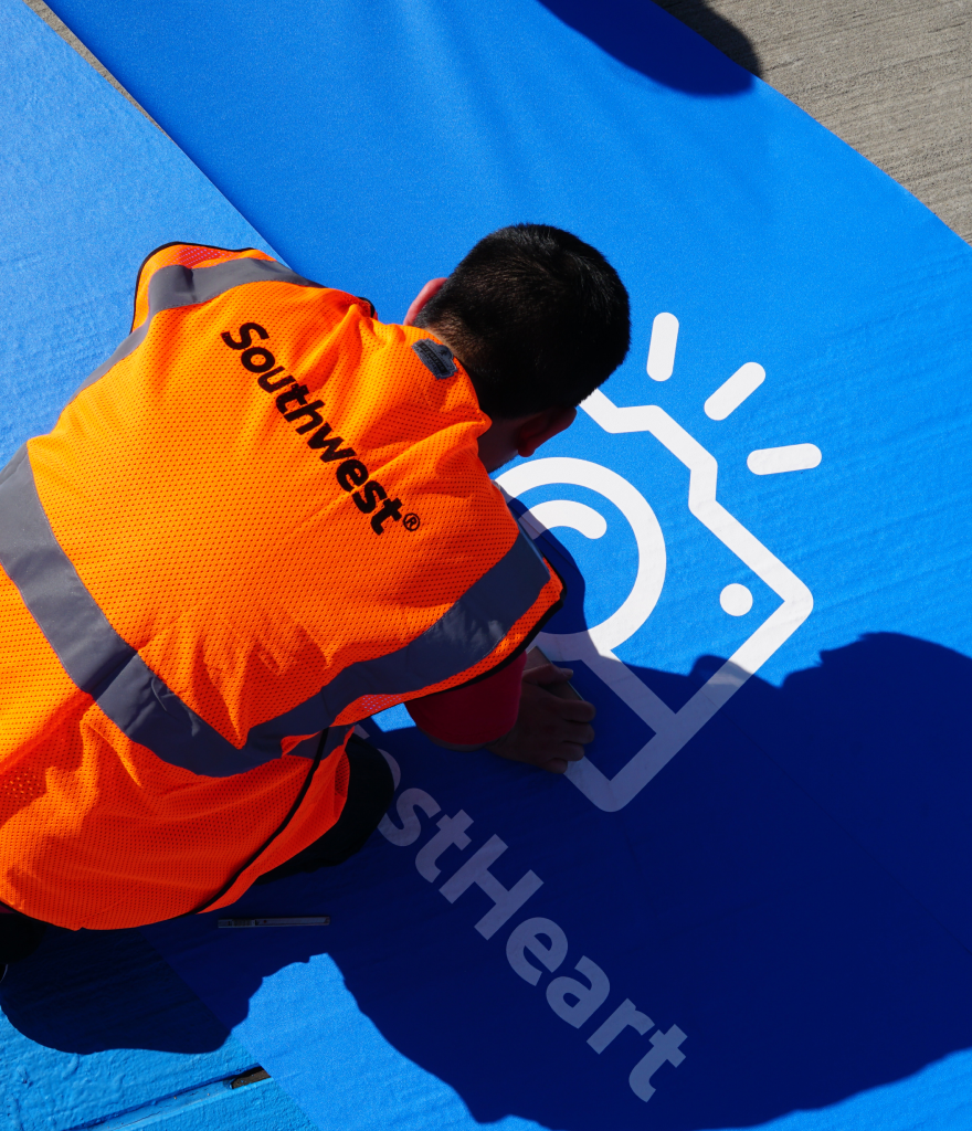 Person in an orange vest with 'Southwest' written on it, kneeling on a blue surface, painting or drawing a white logo or symbol that includes a camera and sun icon.