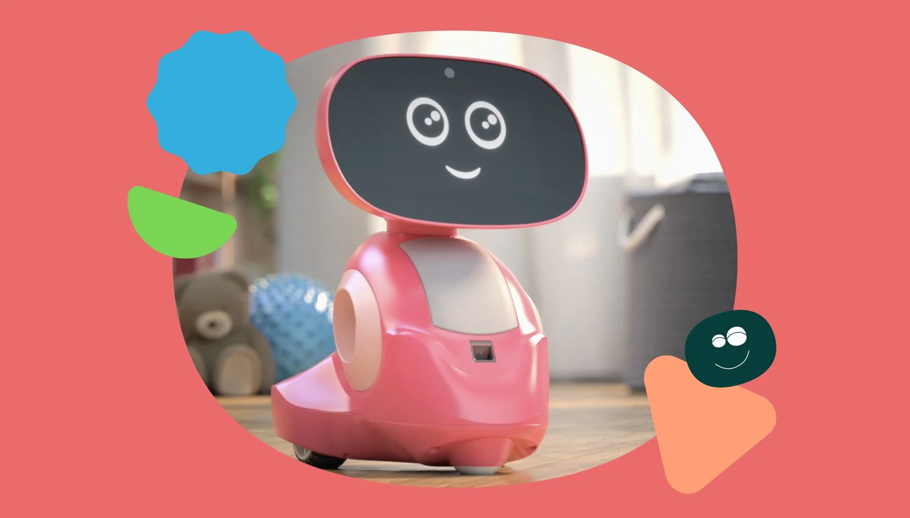 A pink robot with a black screen on its face displaying cartoon eyes and a smiling mouth, situated indoors on a wooden surface with toys in the background.
