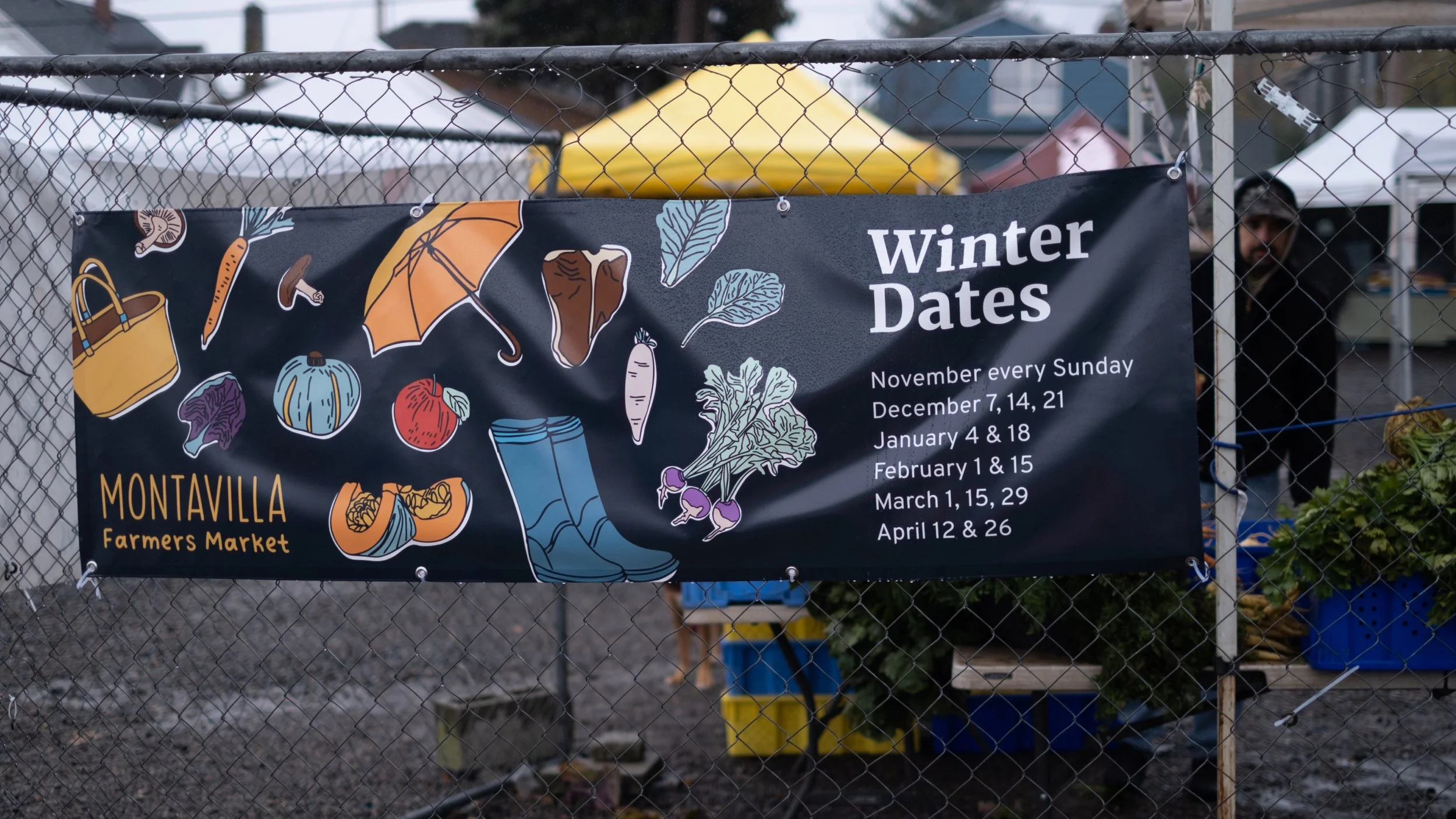 A banner for Montavilla Farmers Market listing winter dates with illustrations of fall vegetables and accessories, attached to a chain-link fence at an outdoor market.