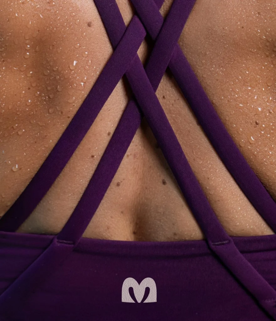 Close-up of a person's back in a purple sports bra with water droplets on skin and purple straps with an "M"-shaped logo.