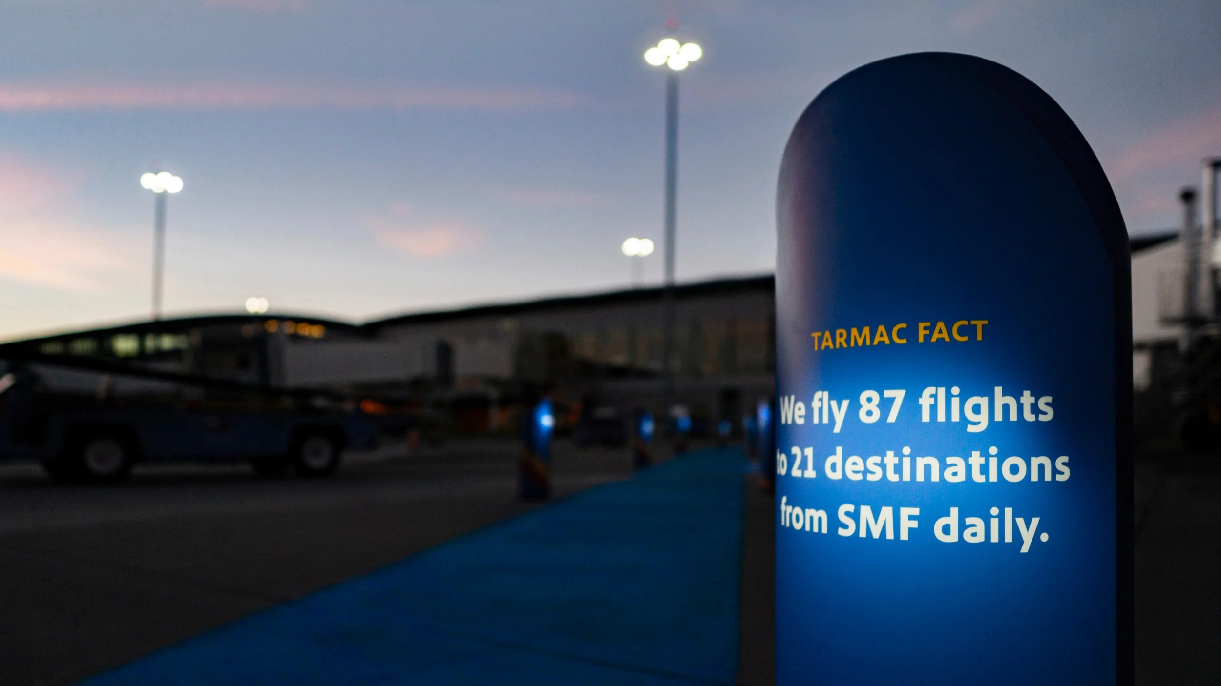 Blue informational stanchion at an airport indicating TARMAC FACT: We fly 87 flights to 21 destinations from SMF daily, against a blurred background of airplanes, vehicles, and airport lights during dusk.
