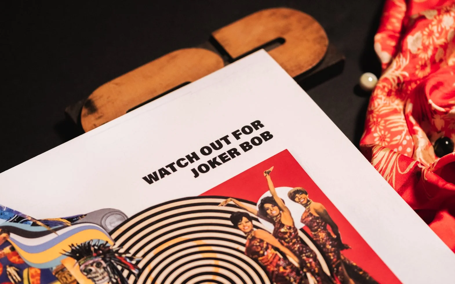 A poster titled 'WATCH OUT FOR JOKER BOB' with an image of women dressed in 70s style clothing, standing in a striped spiral background.