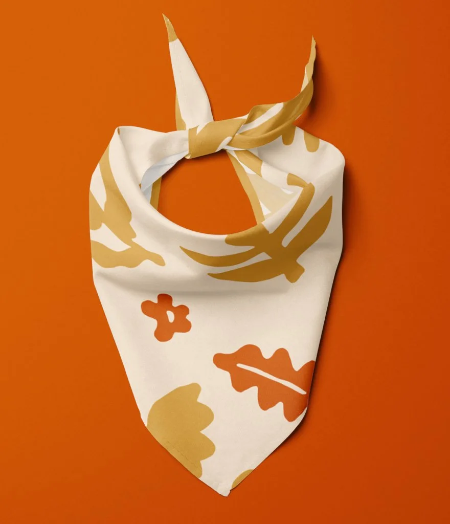 An orange background with an off-white bandana decorated with gold and orange leaf and floral patterns, tied in a knot.