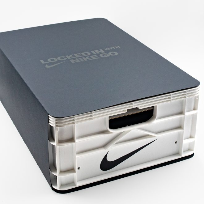 A Nike shoebox with the lid slightly open, showing a white shoe box inside with a black Nike swoosh logo on the front. The top of the box has the text "LOCKED IN WITH NIKE GO".