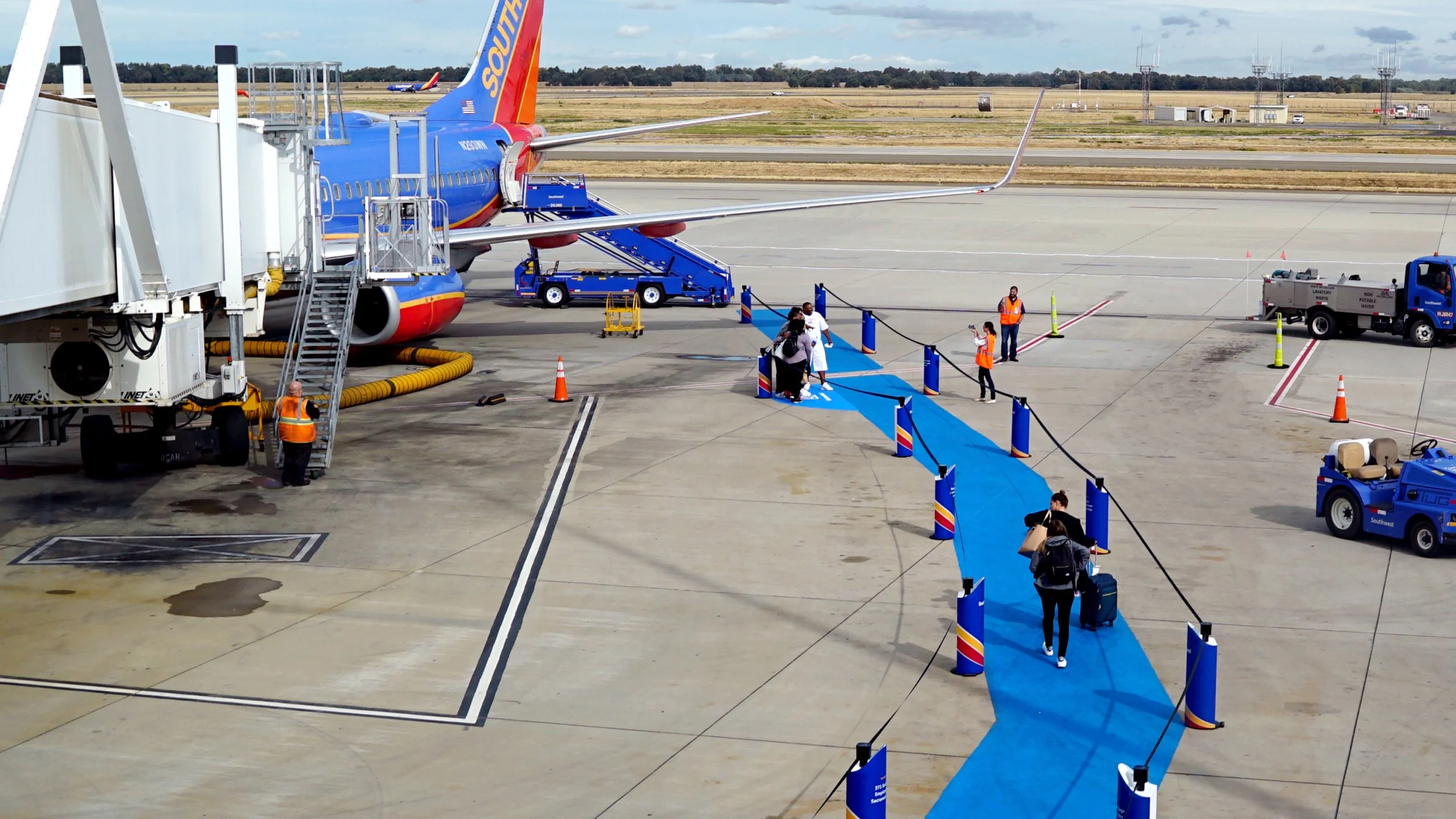 Passengers walking on a blue-painted walkway to board a Southwest Airlines plane at an airport gate, with ground crew members and airport vehicles nearby.