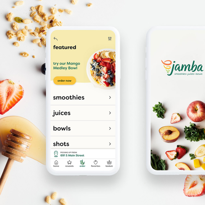 Smartphone displaying a food ordering app with options for smoothies, juices, bowls, and shots, placed on a white surface with fresh strawberries, a honey dipper, and granola scattered nearby.